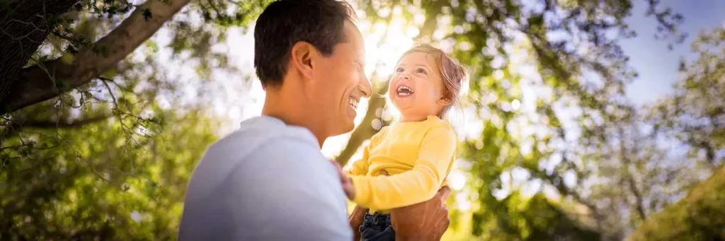 UCHealth Today "raising healthy happy children". Father and daughter laughing under a tree in the park.