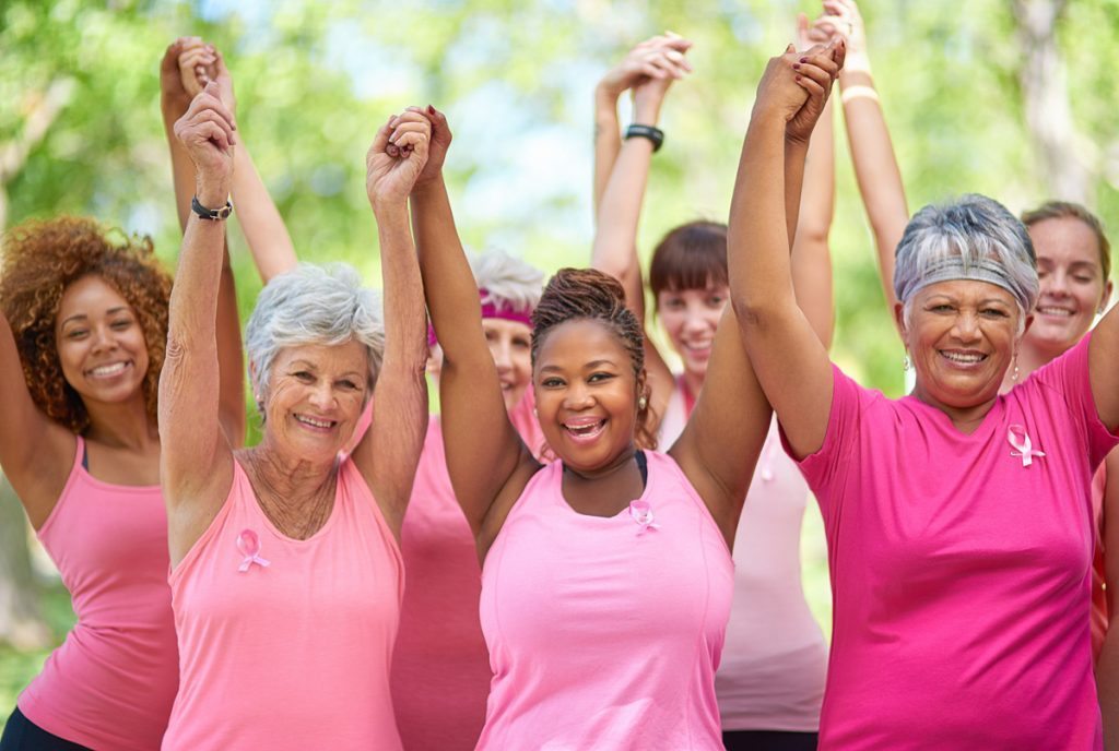 Women in pink shirts, representing breast cancer awareness, hold their hands up in the air.