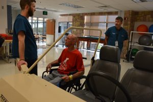 Patrick Buxton, a patient in the Rehabilitation Patient Care Unit at Memorial Hospital Central, works with Andrew Bettlach, an occupational therapist, to practice transferring from a wheelchair into a simulated car. Gregory Braeger, therapy manager, looks on.