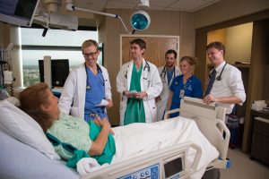 A group of physicians and providers talk with a patient who is sitting up in bed.
