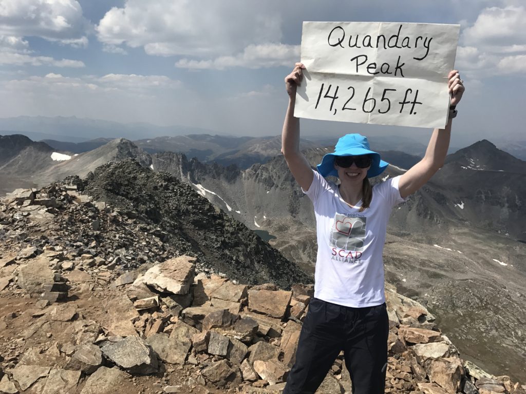 Becky Pomerleau holding a sign saying Quandary Peak 14,265 feet at the top of that peak.