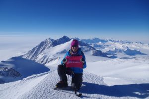 Kim Hess holds a sign that says "UCHealth" as she kneels in the snow with Mount Vinson in the background. Wild swings in temperatures were among the challenges of climbing 'Seven Summits.'
