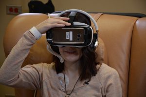 Hannah sits in a chair during a treatment. She wears virtual reality goggles so she can travel virtually while undergoing medical treatments.