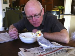 Patient Sonny Hutchison sits at his kitchen table, eating, while his pet bird sits on the table nearby.