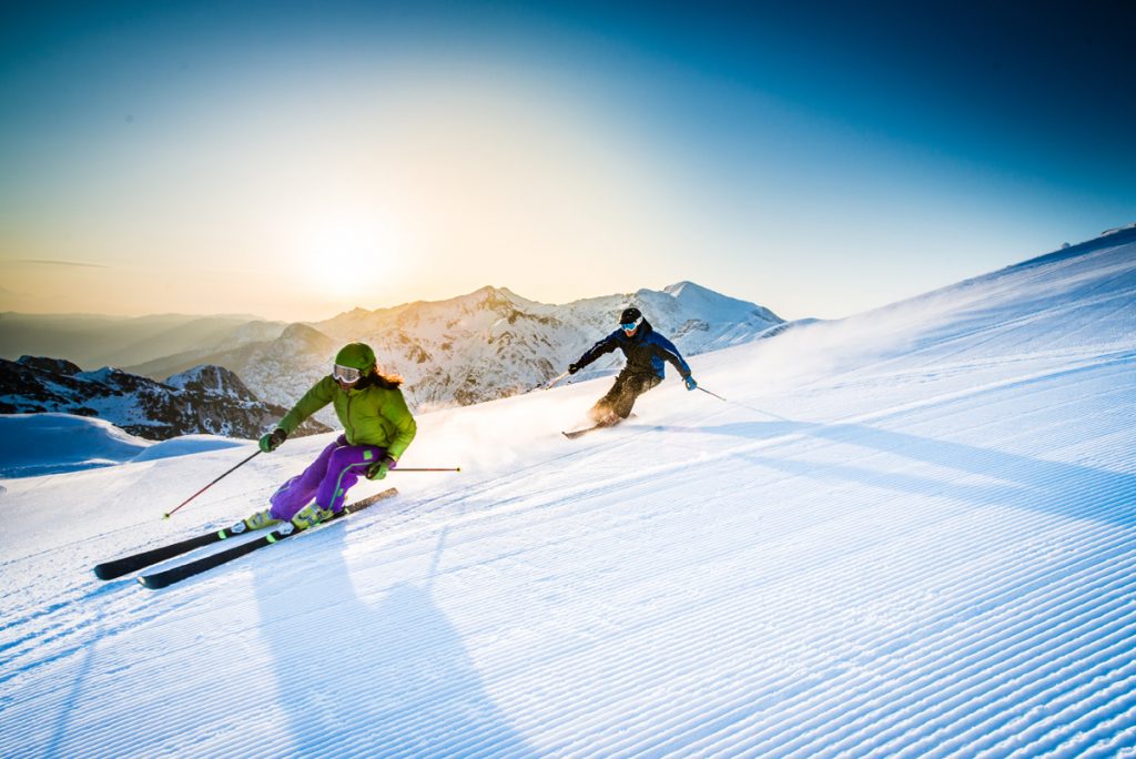 Two skiers ski down a snowy slope with sun in the background.
