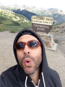 Actor Jordan Leigh in a selfie at the top of Loveland Pass in Colorado with a sign behind him that says Loveland Pass, Elevation, 11,990, Continental Divide.