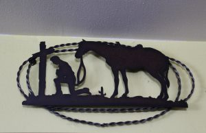 A decorative art piece made of metal, depicting a soldier kneeling at a cross, with his horse nearby.
