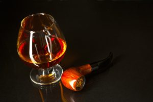 Whiskey glass and smoking pipe on black background. Cognac glass. Brandy glassful. Cognac france. Scotch drink. Smoking pipe