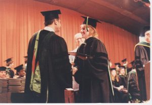 Dr. Jeanne Lewis, wearing a black cap and gown, receives her diploma on a stage at her medical school graduation.