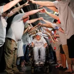 Cancer survivor supporters form a tunnel by lining both sides of a hallway and holding hands up. The survivors walk and wheel beneath their hands toward the Colorado Eagles locker room.
