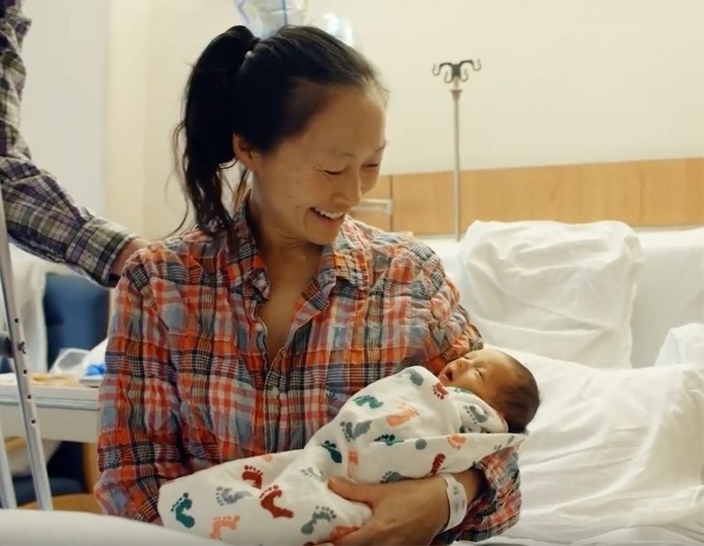 A mother wearing a flannel shirt gazes down at her newborn, who is wrapped in a blanket.