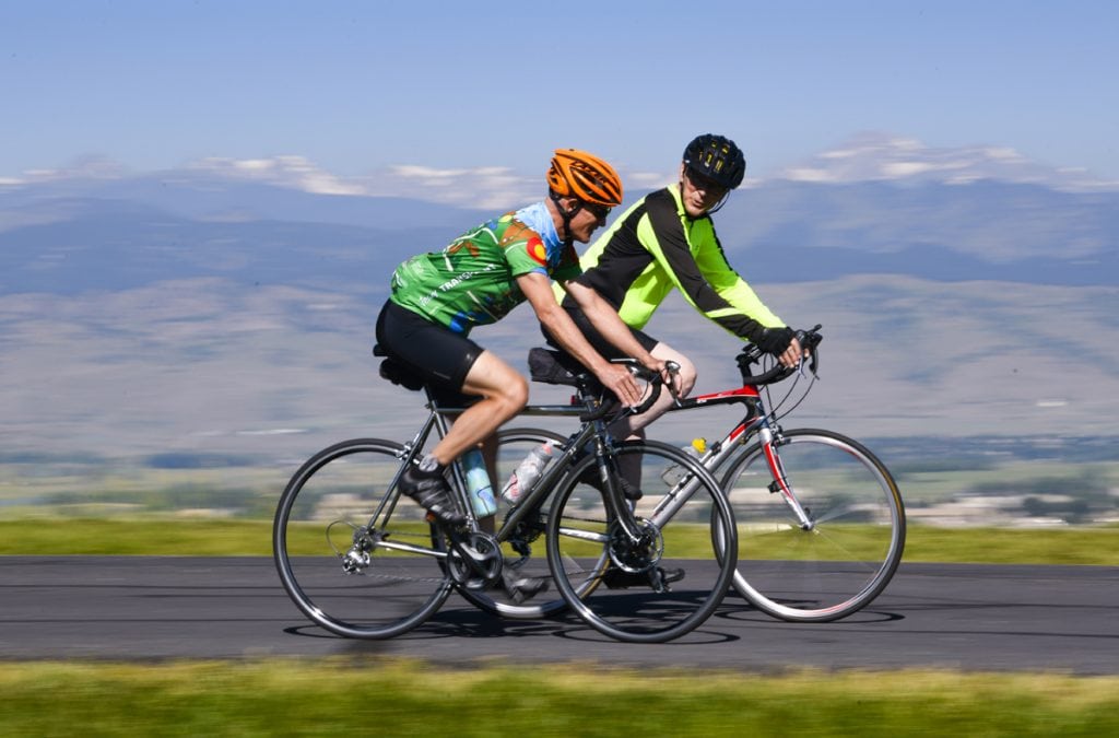 Scott La Point and Jim Eastman cycle together on a road with mountains in the background. They are near Niwot, CO. The men both survived traumatic brain injury, then La Point donated his kidney to Eastman. The men will be cycling in the Transplant Games of America.
