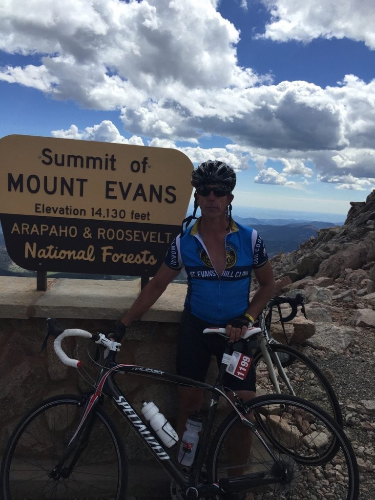 Avid cyclist Rich Trostel is back on his bike thanks to a partial knee replacement. In this photo, he poses at the top of Mt. Evans. Behind him is a sign showing the altitude of the Colorado 14er.