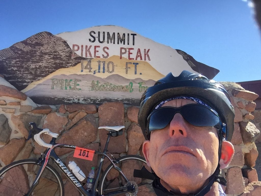 Cyclist Rich Trostel poses in a selfie on the summit of Pikes Peak. His bike and a sign at the summit are behind him.