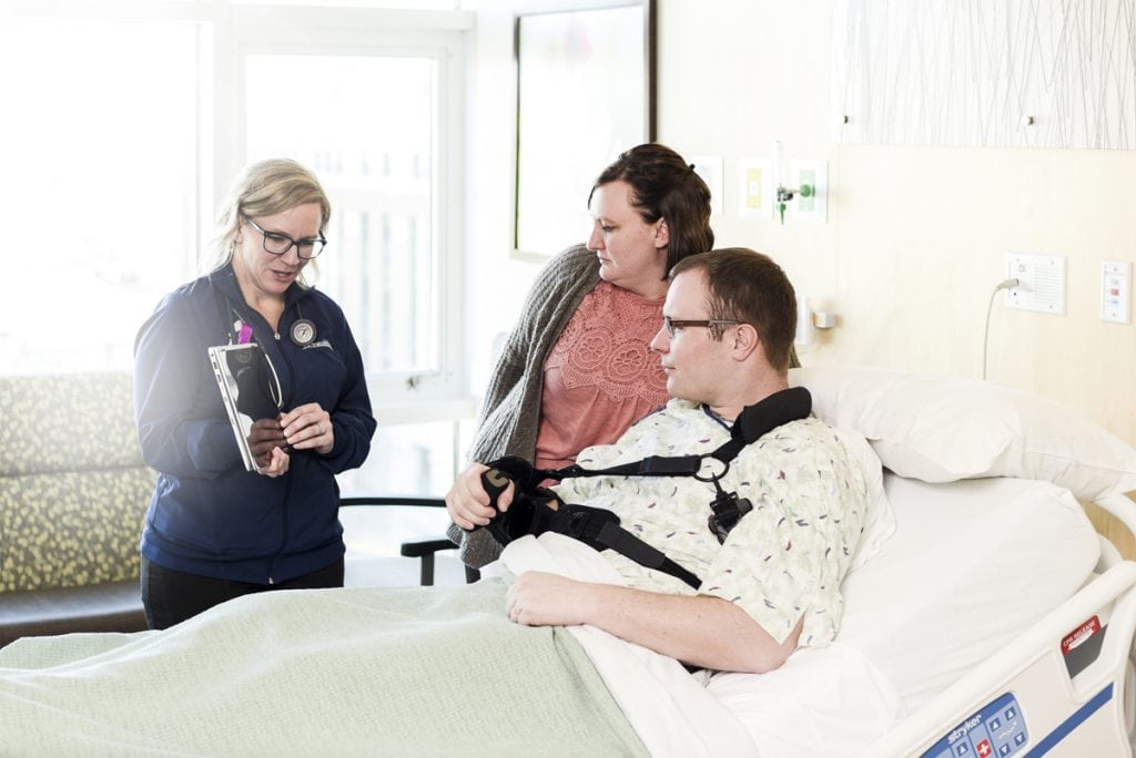 A patient at UCHealth University of Colorado Hospital sits in bed with his wife at his side as a nurse reviews paperwork with him.