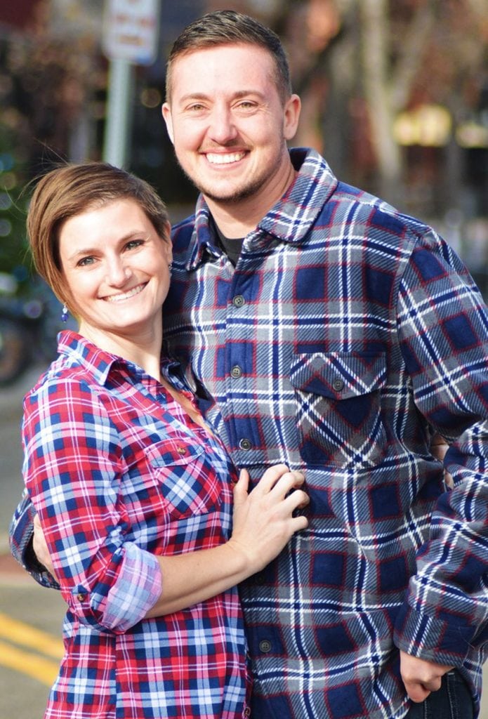 A copule poses for a photo. They were flannel shirts. On the left is a woman. On the right is Mal Foust, a transgender male. He's got a scruffy beard and is much taller than his partner.