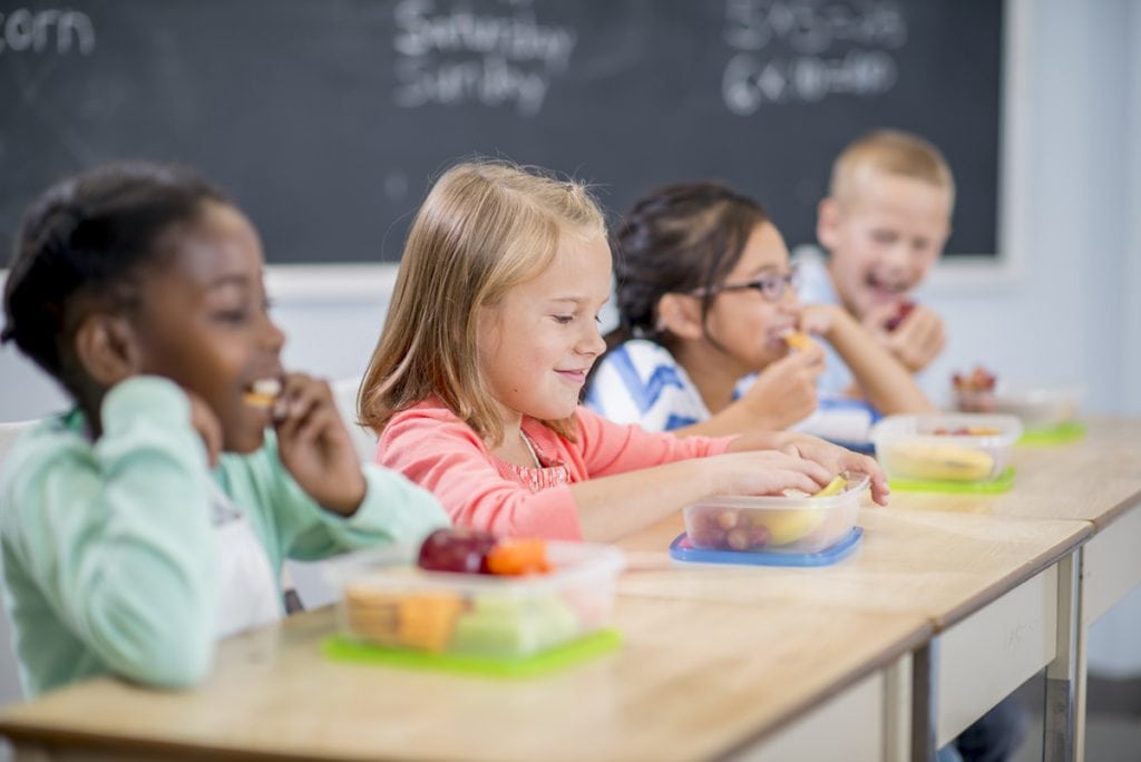 A group of children sit at their desks in a classroom eating lunch.