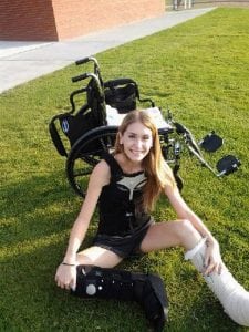 A young woman sits in grass with her wheel chair behind her.