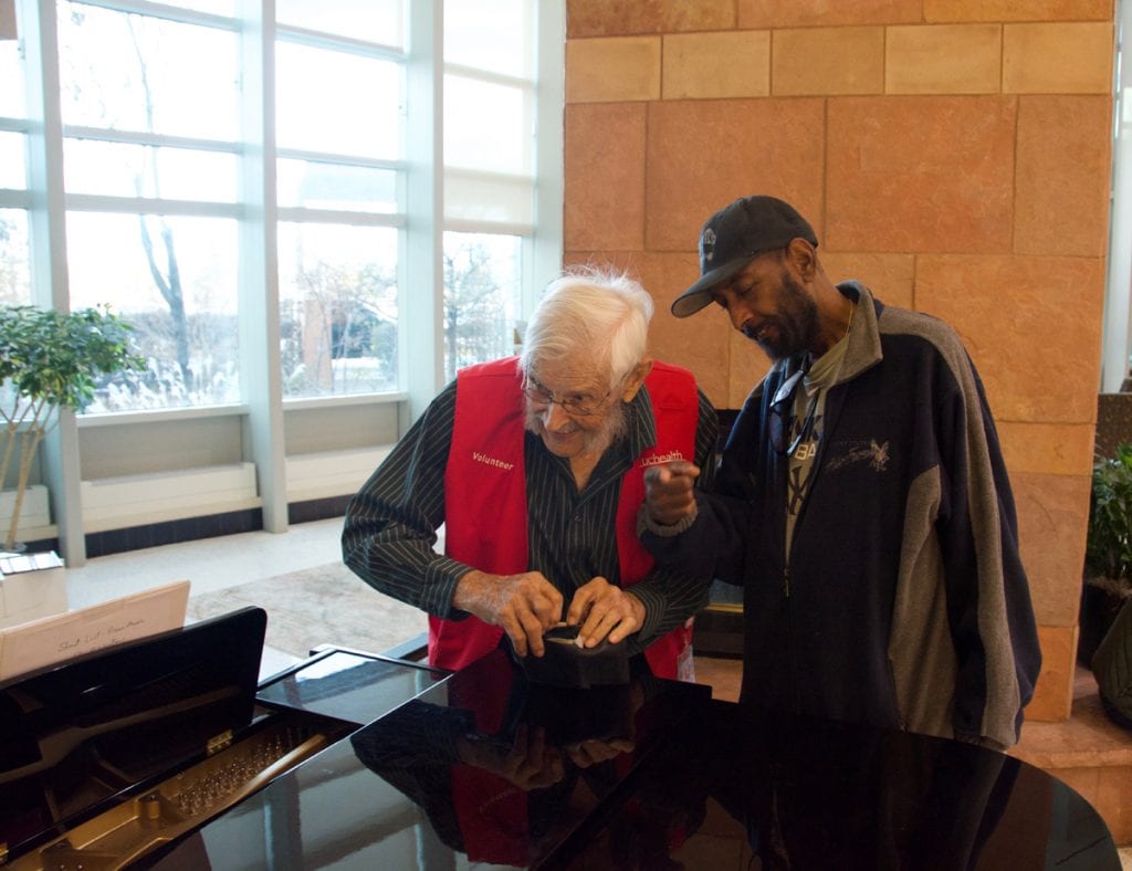 Two men smile as they lean over a piano and sing.