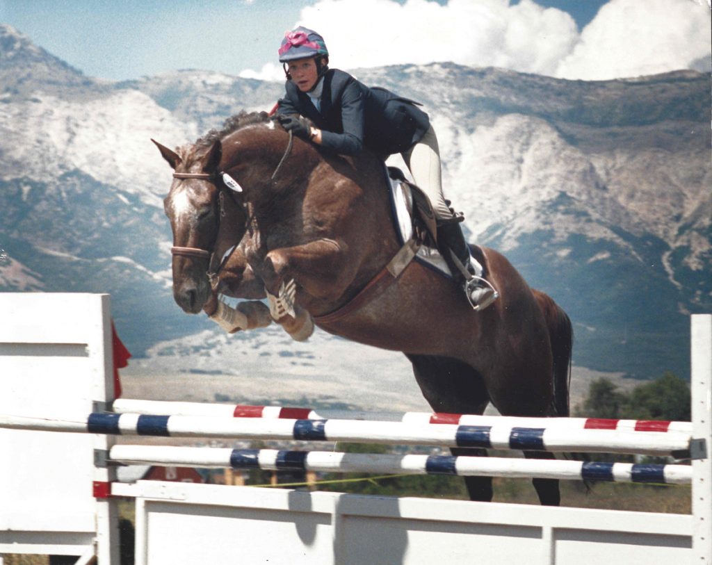 A young woman jumps on her horse during an equestrian competition with mountains in the background.