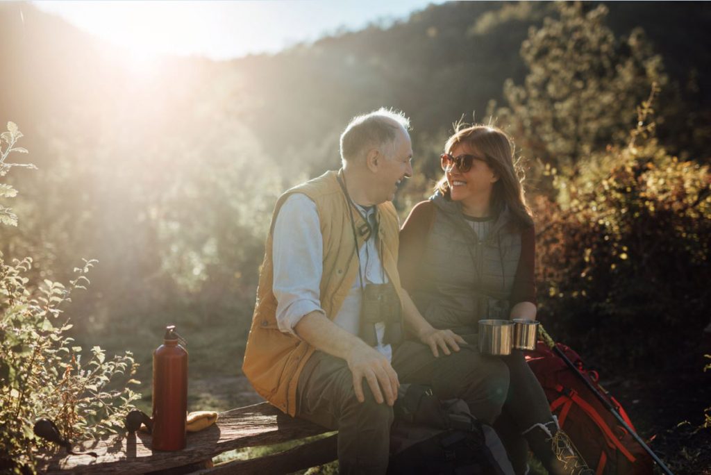 An older couple takes a break from hiking. They have binoculars and water bottles.