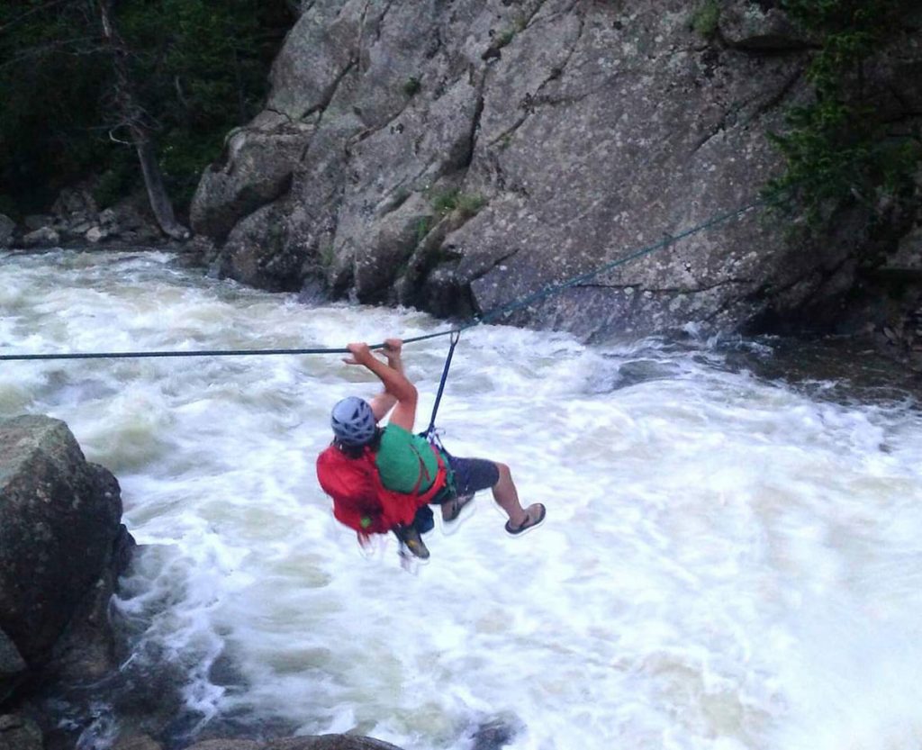 A man crosses a white water on a churning river. He's sliding on a rope to get across.