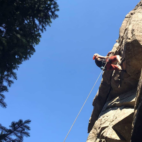 a man is rock climbing outdoors. We see him from below with a big cliff, pine trees and blue sky.