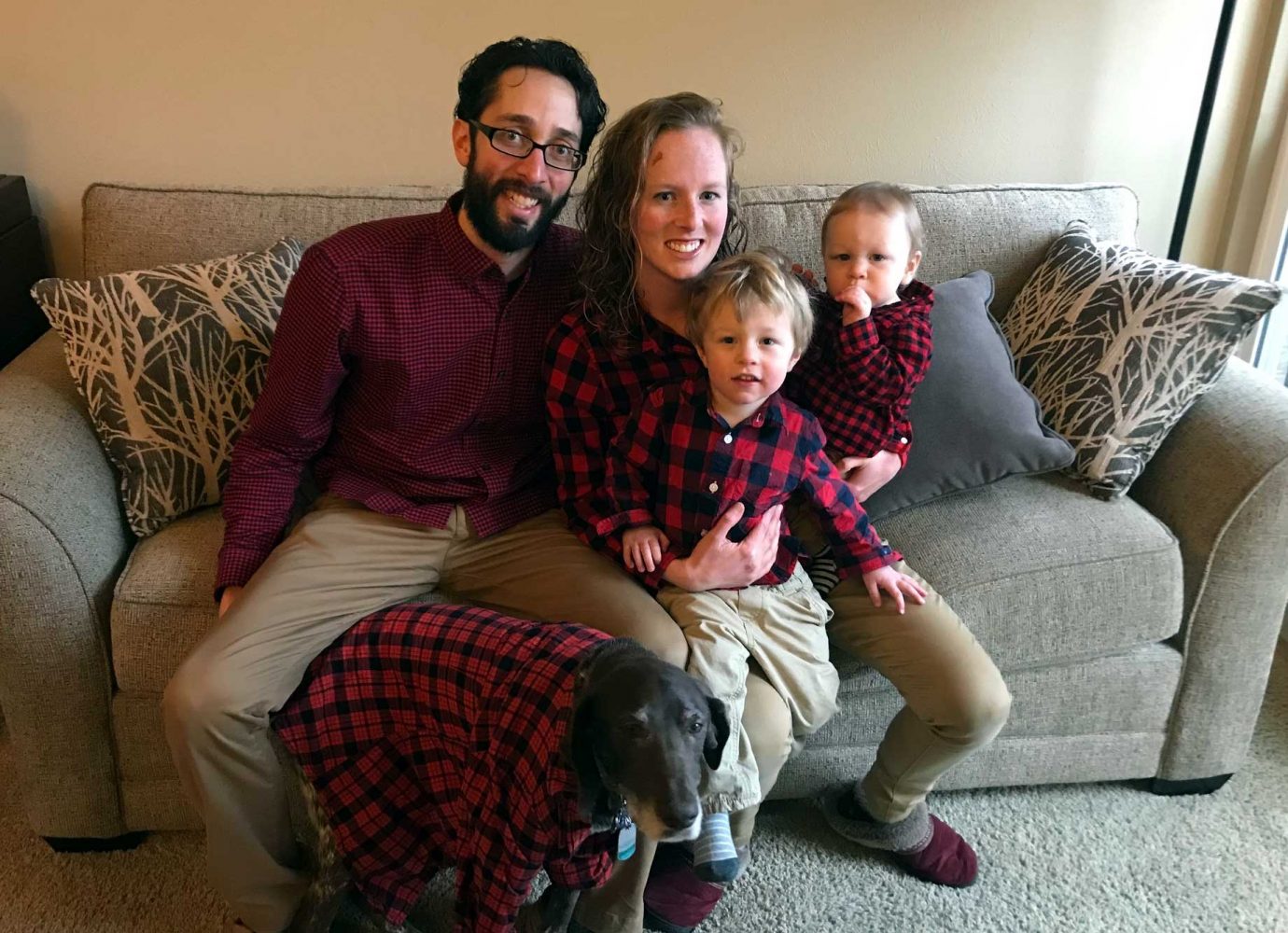 dad, mom, two young kids and a dog sitting on the couch wearing red flannel.