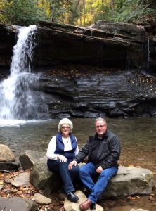 An older couple poses with a small waterfall behind them.