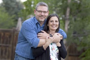 husband with arms around wife, both looking at camera