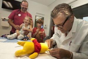 Doctor sews up a stuffed bear while two kids watch in an emergency room.
