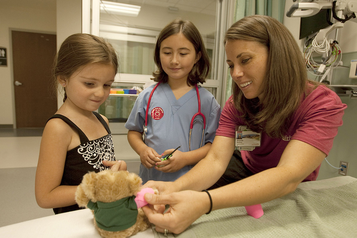 A nurse bandages up a stuffed bear as two young girls watch.