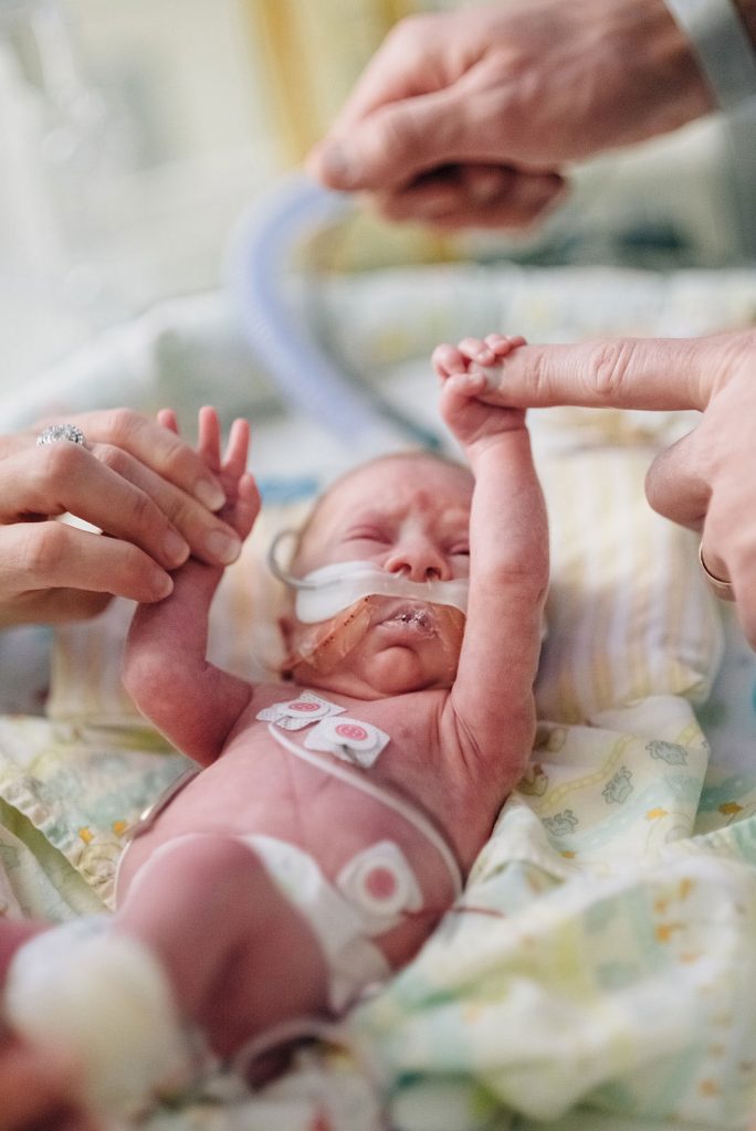 A preemie baby grasps onto his parents' hands.