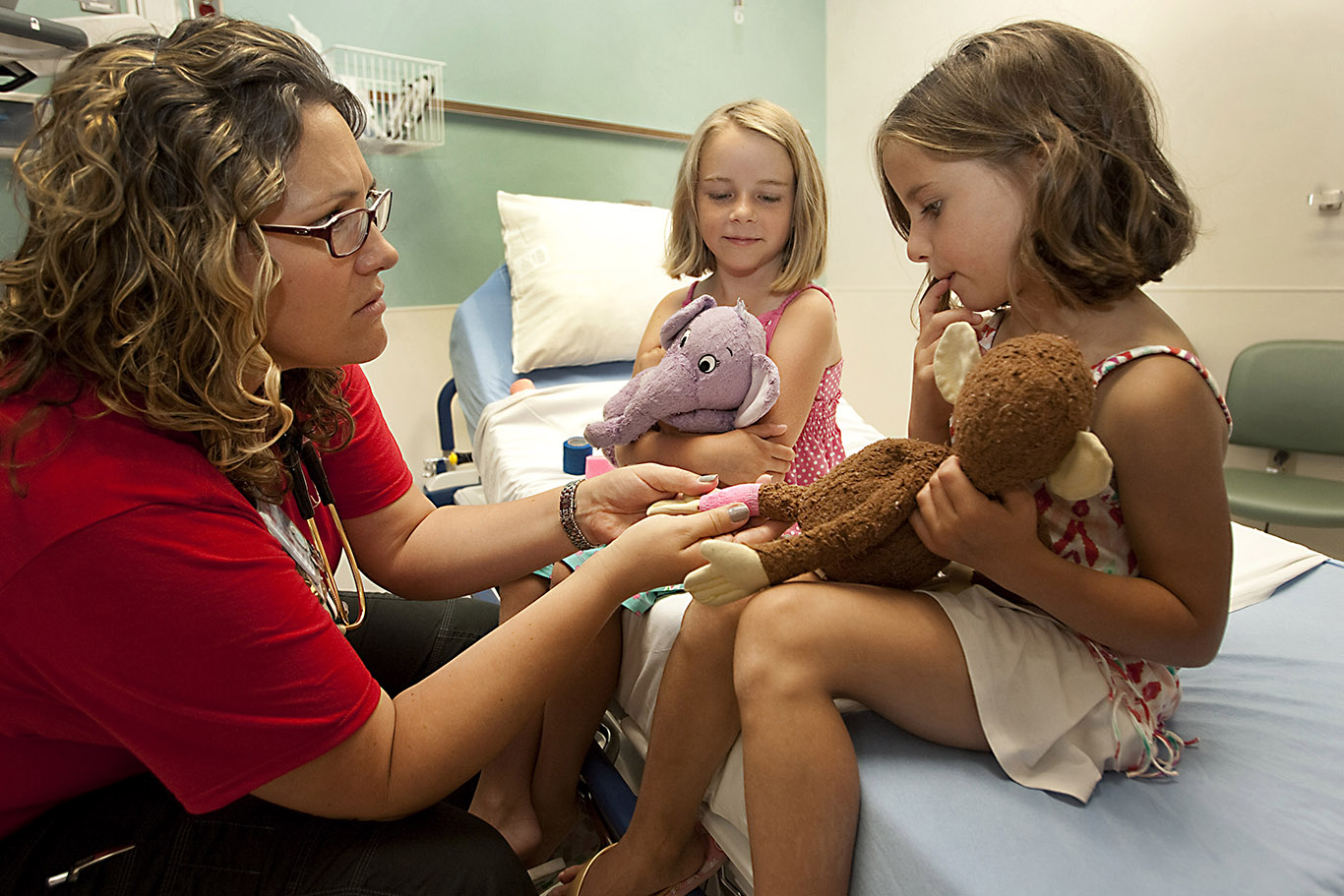 Two girls hug their stuffed animals as a nurse pretends to tend to the stuffed animals' injuries.
