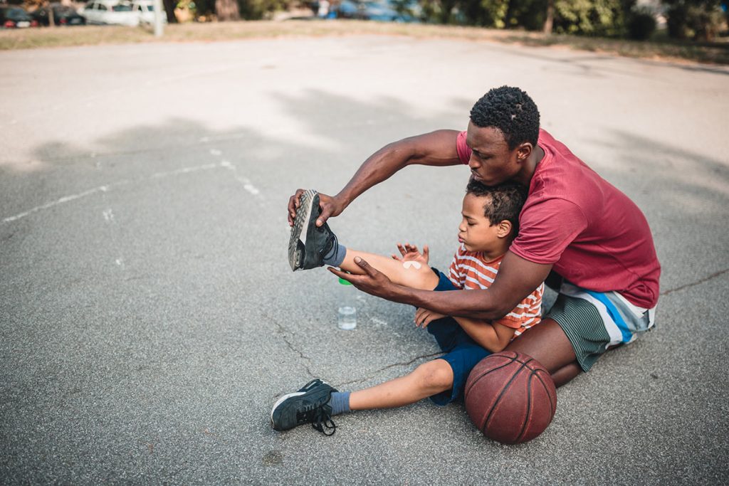 A father checks his son's ankle while sitting on the ground playing basketball outside.