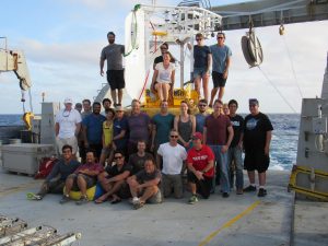 about 20 crew members stand on a massive ship in the middle of the ocean for a photo