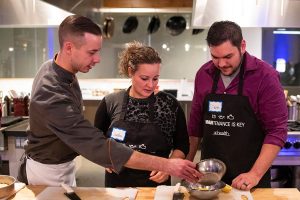 Three people stand over cutting boards in a kitchen, preparing food.