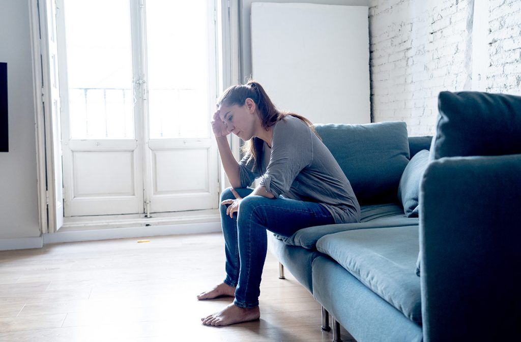 A woman is sitting on a couch with forehead in hand. she is feeling blue.