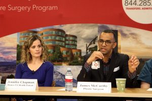 Doctor speaks in a panel-like presentation while his former patient sits next to him and listens. The patient had returned to thank hospital staff for her care.