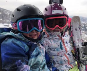 Two young girls are shown in ski gear, including helmets and goggles, in this photo.