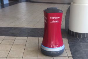 A red, cylindical robot on wheels moves through a hallway to deliver medications