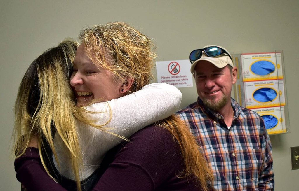 Keith's wife hugs the urgent care doctor who diagnosed her husband with colon cancer months earlier. keith is in the background smiling.