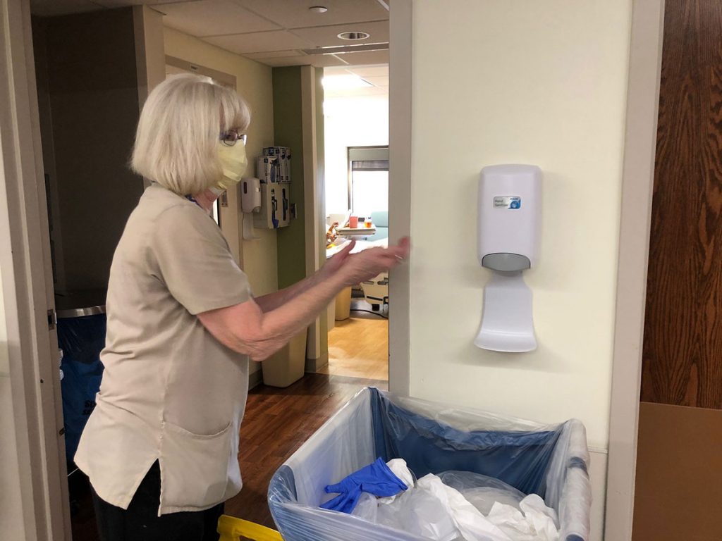 hospital housekeepers, like this one wearing a mask and reaching for hand sanitizer, are on the front lines during covid pandemic.