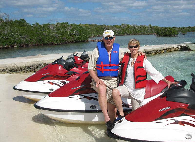 Dave with wife sitting on jetskis on sandy beach.
