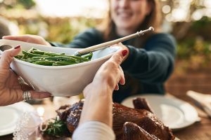 Holiday gatherings in 2020 will need to be smaller and safer, like this celebration outside. A person hands green beans over a turkey at a table outdoors.