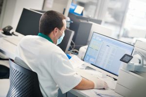 Lab technician sitting at a computer. A major data set called COVIDome aims to identify specific patterns – changes in proteins, red blood cells or immune response, for example – that can help answer the question: Why does COVID-19 affect people differently?