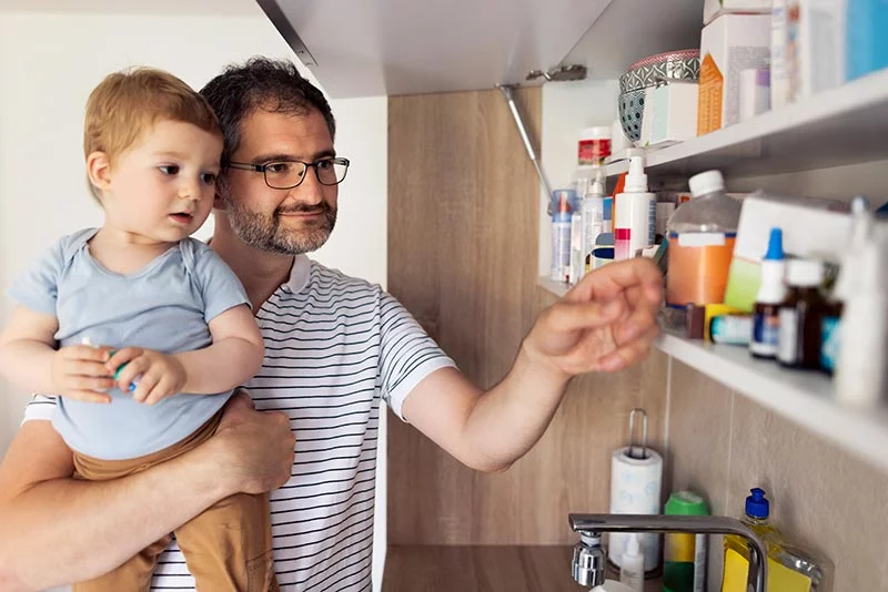 Man holding his son and looking into his medicine cabinet for must-haves. Having basic over-the-counter medications and first-aid supplies makes it much easier to handle minor injuries or illnesses. Photo: Getty Images.