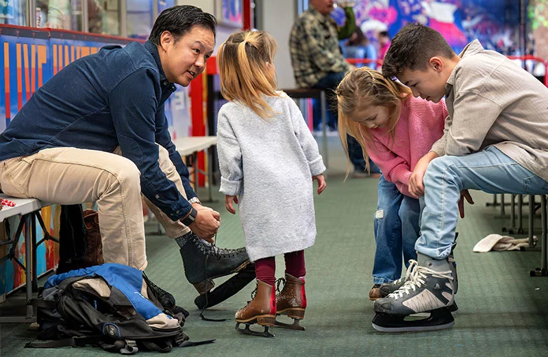 Lee helps his children lace up their skates before they head out on the ice in Pueblo. From left to right, the children are Collins, Calihan and Cruz. Calihan had been asking her parents for skating lessons, and Collins wanted to take her rental skates home with her. Photo by Mike Sweeney, for UCHealth.