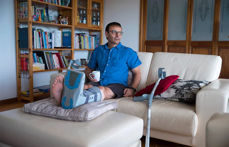 man on his couch with coffee, with crutches nearby and a foot brace on. Preparing your home and support system before surgery can help you recover safely and reduce stress during healing. Photo: Getty Images.